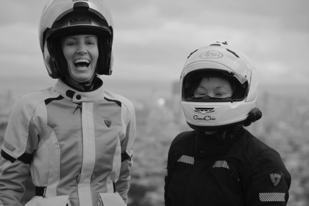 Women Who Ride Motorcycles: Cristi Farrell and Joanne Donn - co-hosts of the Moterrific podcast. The image shows a white woman and an Asian woman wearing motorcycle jackets and helmets. They are laughing.