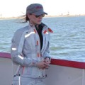 Motorcyclist Cristi Farrell looking out at the water. She is dressed in a RevIt jacket, a hat and sunglasses.
