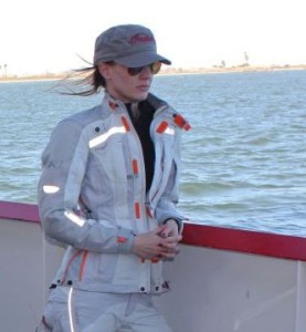 Motorcyclist Cristi Farrell looking out at the water. She is dressed in a RevIt jacket, a hat and sunglasses.