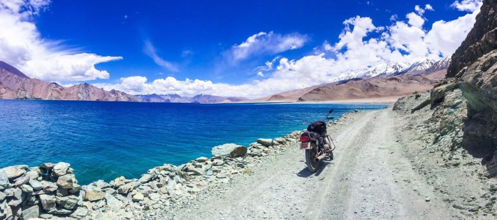 Riding on the banks of Pangong Tso approaching Chusul in Ladakh (Image shows a motorcycle parked on a gravel road bordering a right blue lake with mountains and cloud skies in the distance)