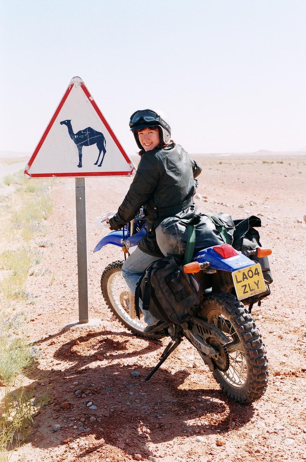 Women Who Ride: Lois Pryce in Morocco next to a road sign depicting a camel