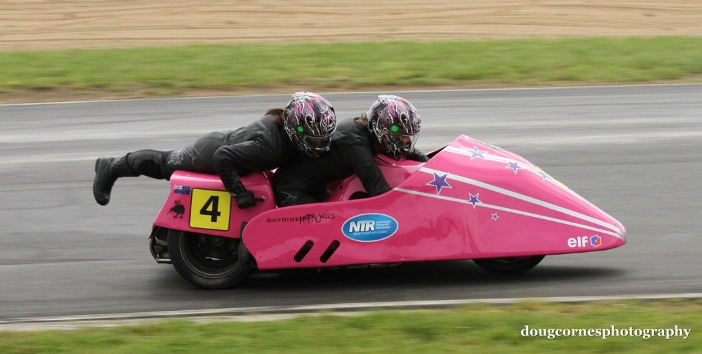Women Who Ride: Tracey Bryan with Tina McKeown on their sidecar "Pinky". The image depicts a pink motorcycle sidecar on a race track with two motorcyclists dressed in black suits riding it.